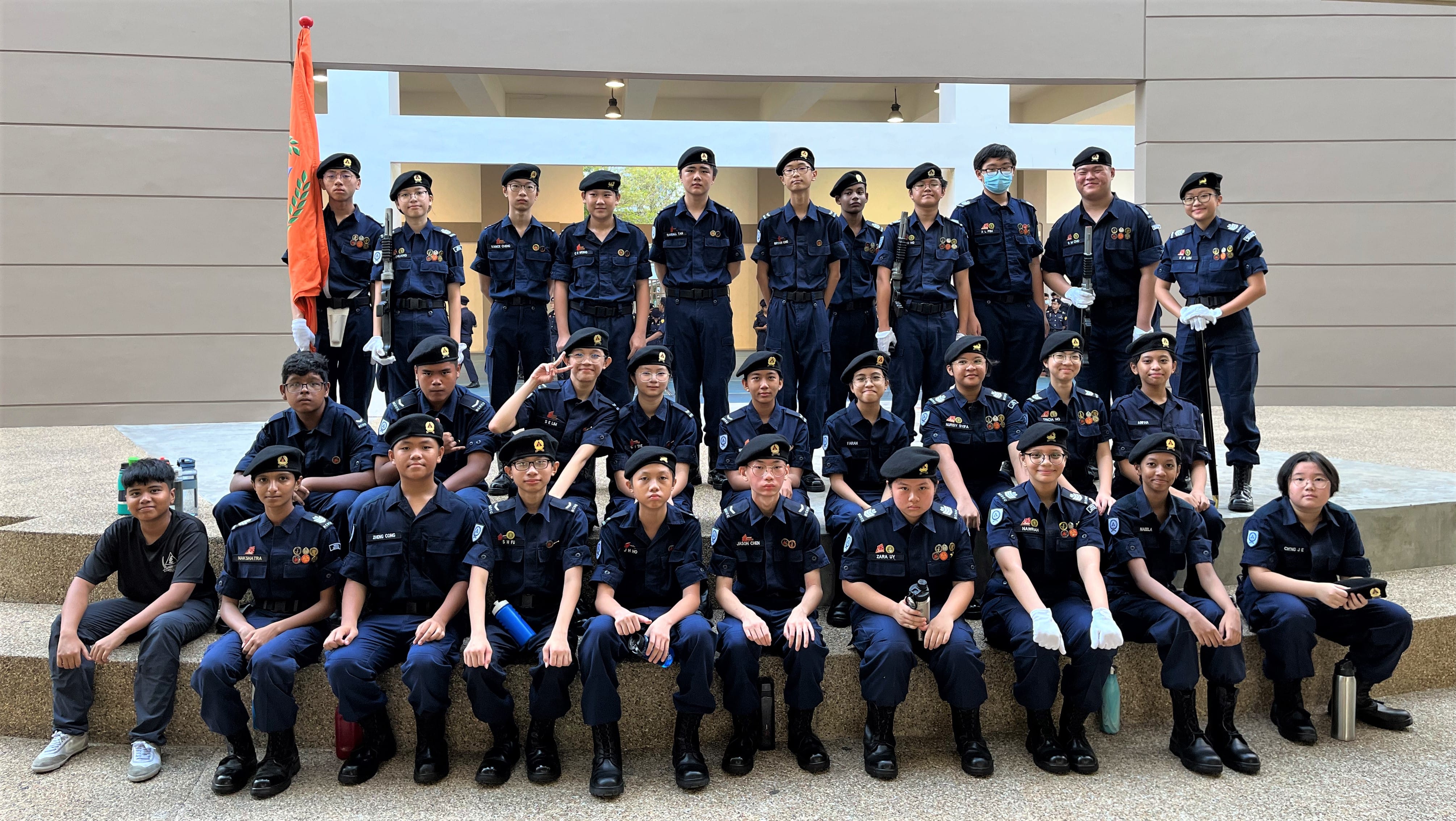 A large group of youth cadets in dark blue uniforms and black berets posing together, with an orange flag visible.